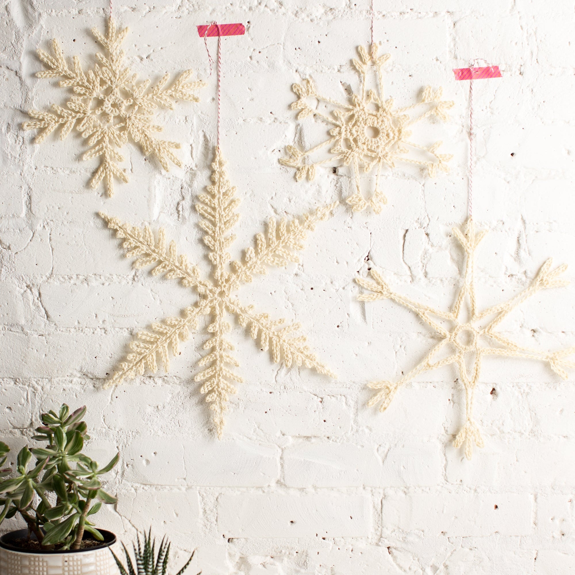 Decorative snowflake ornaments hanging on a white brick wall with two potted plants at the bottom.