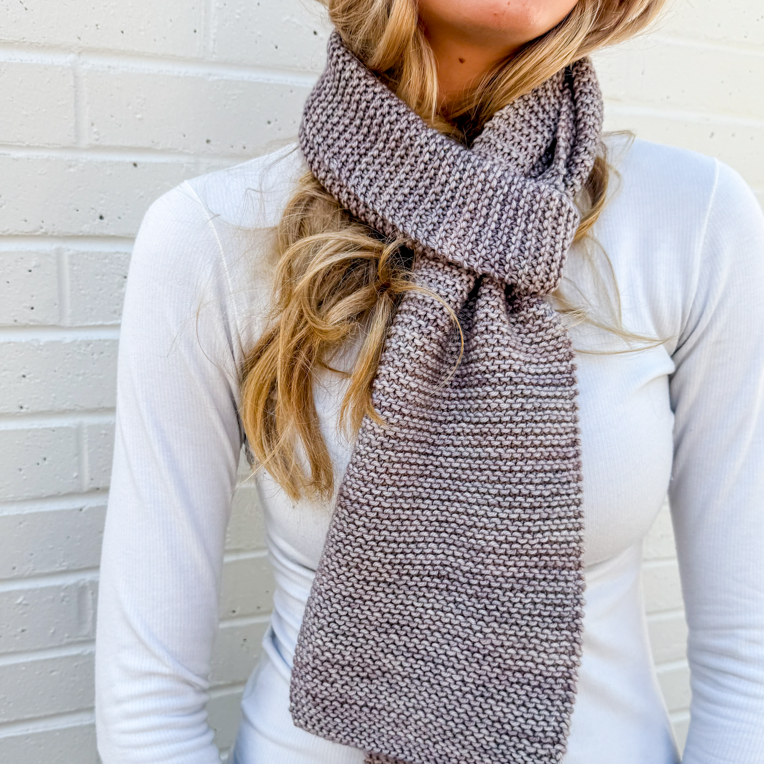 Woman wearing a gray learn how to knit scarf against a light brick wall
