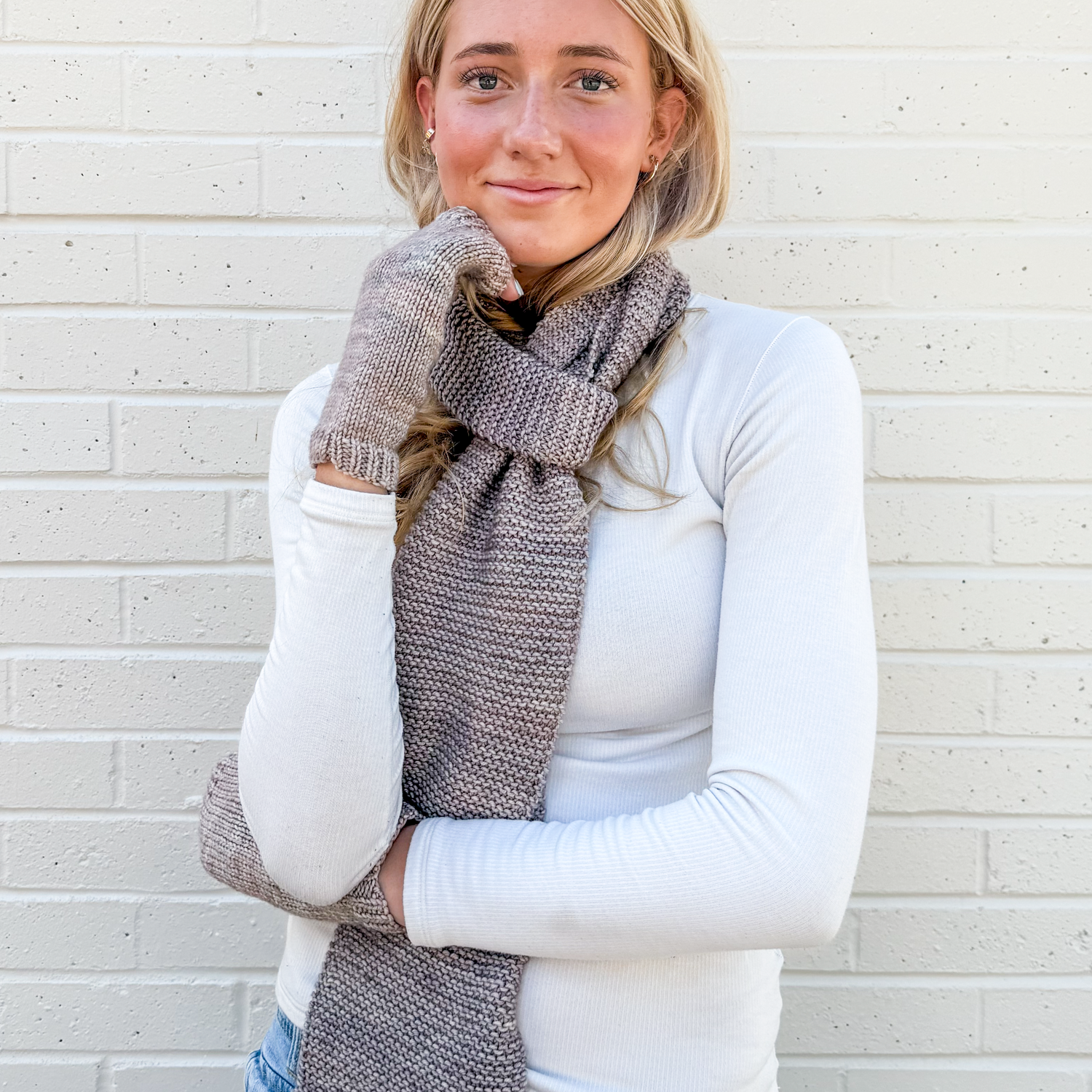 Woman wearing a learn how to knit scarf with matching mitts against a light brick wall
