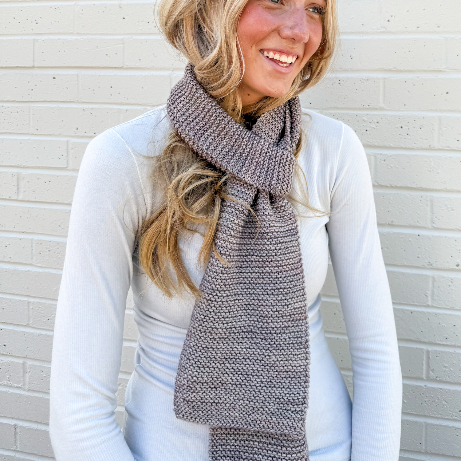 Woman wearing a gray learn how to knit scarf against a light brick wall